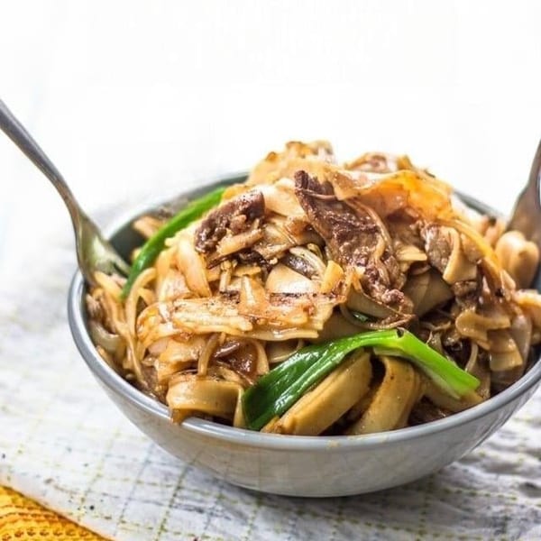 A bowl of stir-fried noodles with beef, mushrooms, and green onions, with a fork.