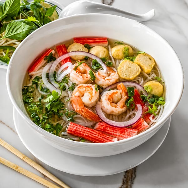A bowl of pho with shrimp, crab sticks, fish cakes, onions, and herbs, with a side of basil.