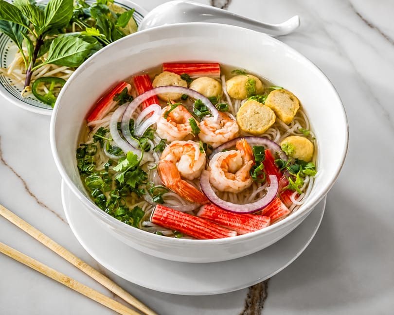 A bowl of pho with shrimp, crab sticks, fish cakes, onions, and herbs, with a side of basil.