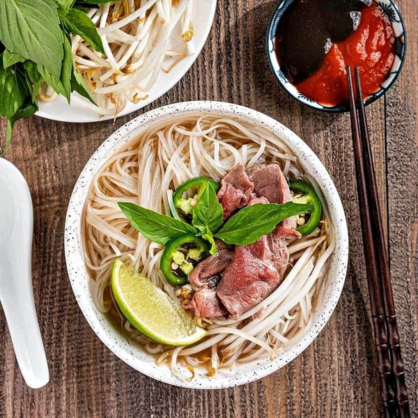 Bowl of pho with beef, noodles, bean sprouts, jalapeños, basil, and lime on a wooden table.