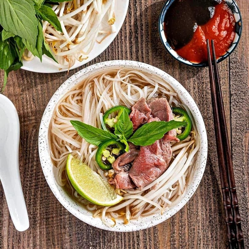 Bowl of pho with beef, noodles, bean sprouts, jalapeños, basil, and lime on a wooden table.