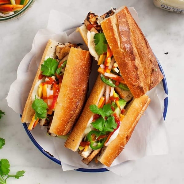 Four banh mi sandwiches with grilled chicken, pickled vegetables, cilantro, and sauces on a blue plate.