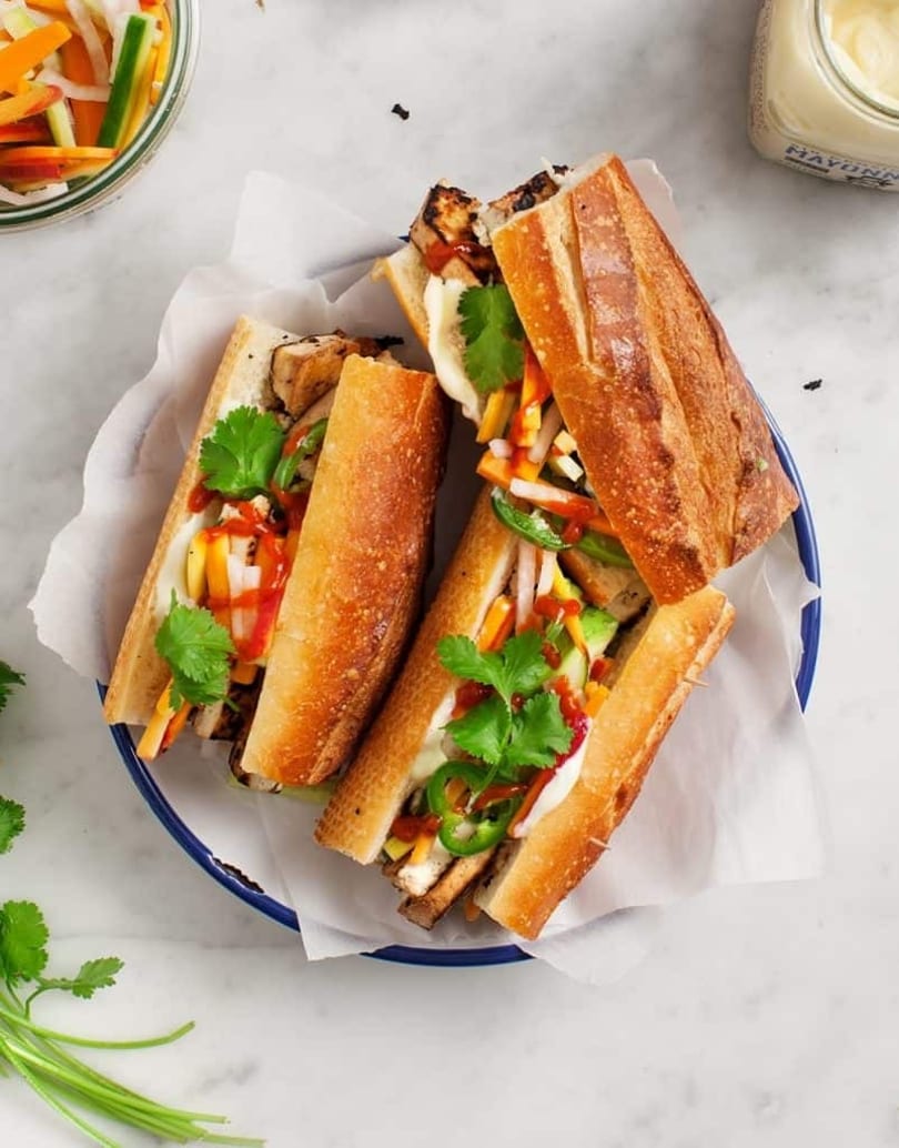 Four banh mi sandwiches with grilled chicken, pickled vegetables, cilantro, and sauces on a blue plate.