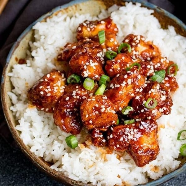 Sesame chicken with white rice, garnished with green onions, in a rustic bowl.